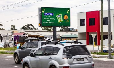 Digital billboard on a roadside promoting Daawat Biryani rice, with the message "This Eid's Biryani, Only Daawat Biryani." Cars are passing by in front of the sign, and buildings are visible in the background.