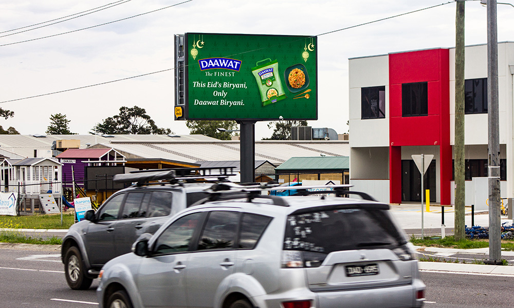 Digital billboard on a roadside promoting Daawat Biryani rice, with the message "This Eid's Biryani, Only Daawat Biryani." Cars are passing by in front of the sign, and buildings are visible in the background.